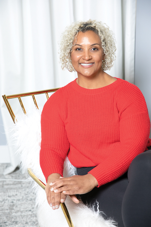 A person with short, curly hair wearing a red sweater sits on a chair in front of white curtains, smiling at the camera.