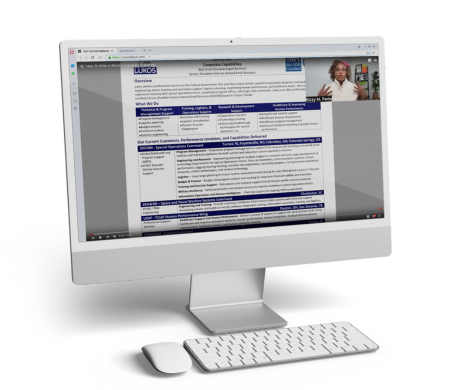 A desktop computer screen displays a document with a detailed table. A small video of a person speaking is situated at the top right corner of the screen. A white keyboard and mouse are in front of the monitor.