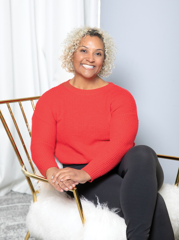 A person with short curly blonde hair smiles while seated, wearing a red sweater.