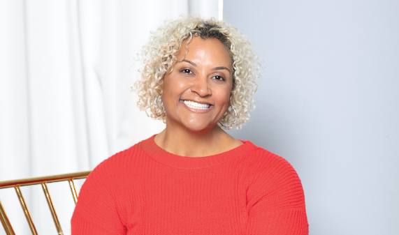 A person with short curly blonde hair smiles while seated, wearing a red sweater.