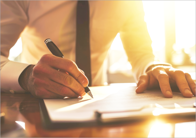 A person in business attire sitting at a desk, holding a pen, and signing a document in a sunlit room.